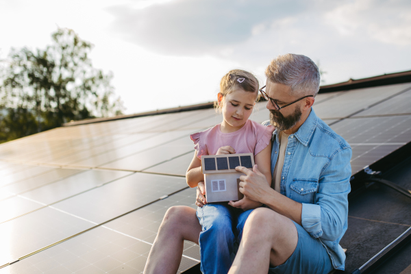 Daughter and father on roof with solar panels, holding model of house with rooftop photovoltaic system.