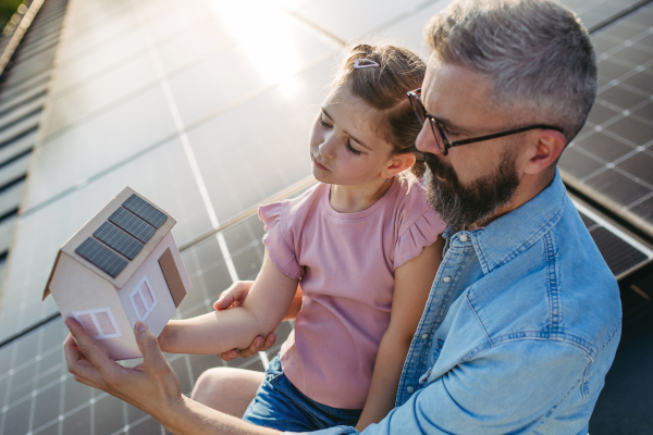 Daughter and father on roof with solar panels, holding model of house with rooftop photovoltaic system.