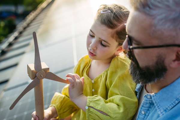 Daughter and father on roof with solar panels, holding model of wind turbine. Rooftop solar or photovoltaic system. Sustainable future for next generation concept.