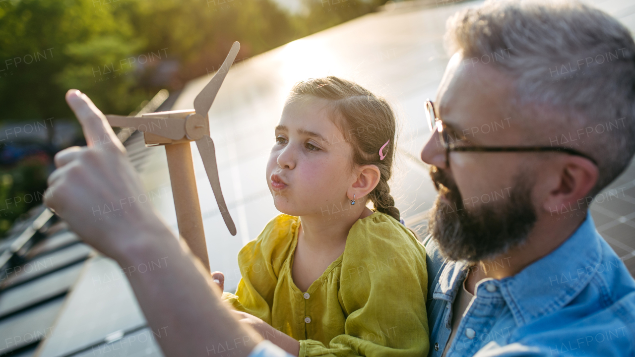 Daughter and father on roof with solar panels, holding model of wind turbine. Rooftop solar or photovoltaic system. Sustainable future for next generation concept.