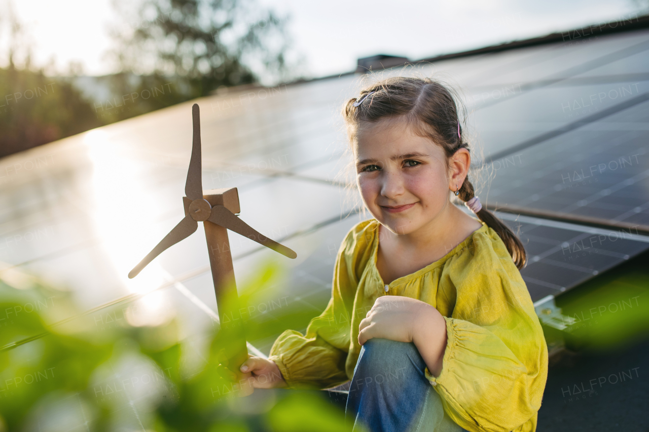 Cute girl on roof with solar panels, holding model of wind turbine. Rooftop solar or photovoltaic system. Sustainable future for next generation concept.