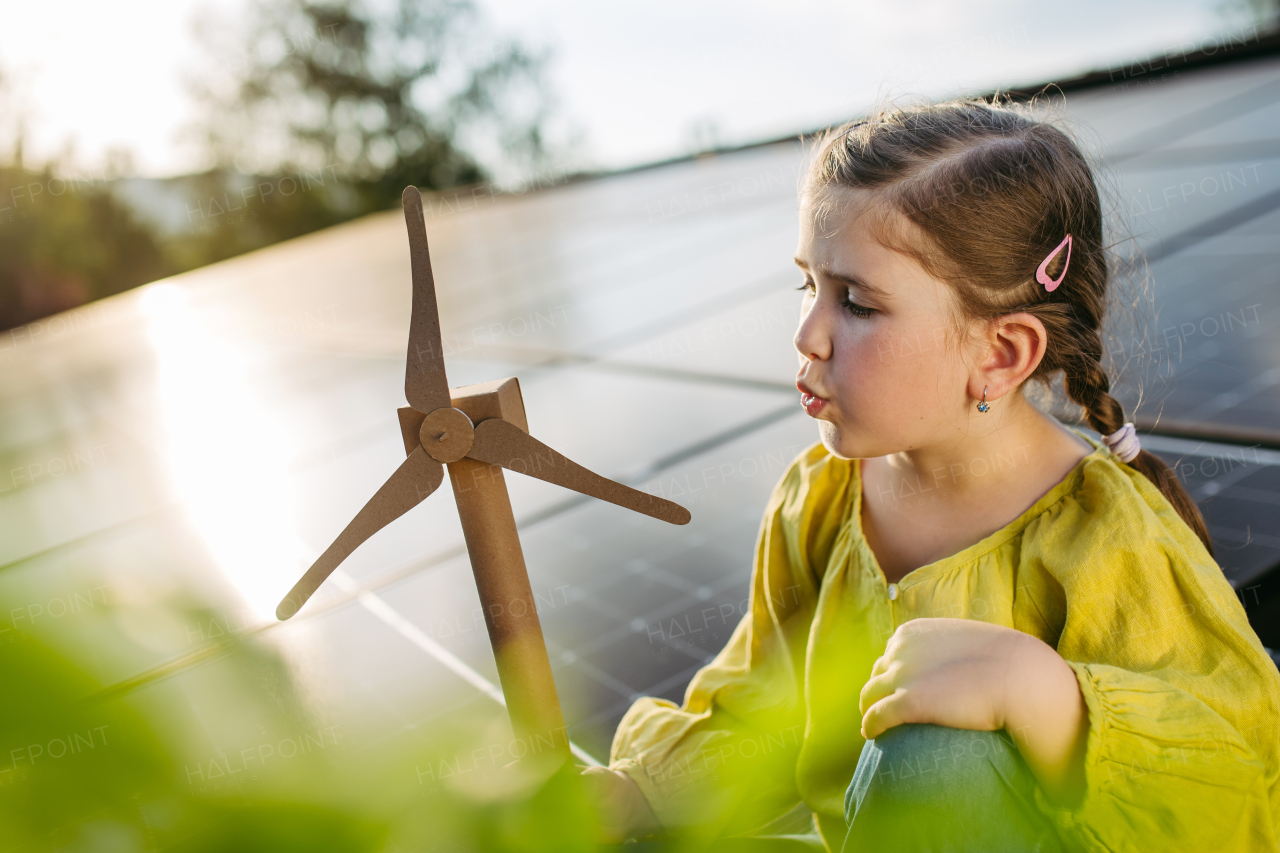 Cute girl on roof with solar panels, blowing into model of wind turbine. Rooftop solar or photovoltaic system. Sustainable future for next generation concept.