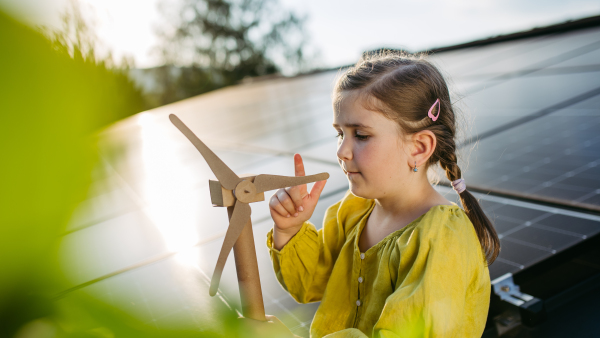 Cute girl on roof with solar panels, spinning blades of wind turbine model. Rooftop solar or photovoltaic system. Sustainable future for next generation concept.