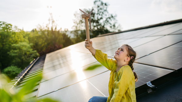 Cute girl on roof with solar panels, holding model of wind turbine. Rooftop solar or photovoltaic system. Sustainable future for next generation concept.