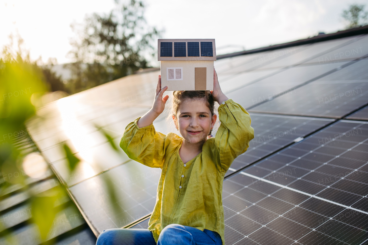 Cute girl holding model of house with solar panels, sitting on rooftop solar or photovoltaic system.