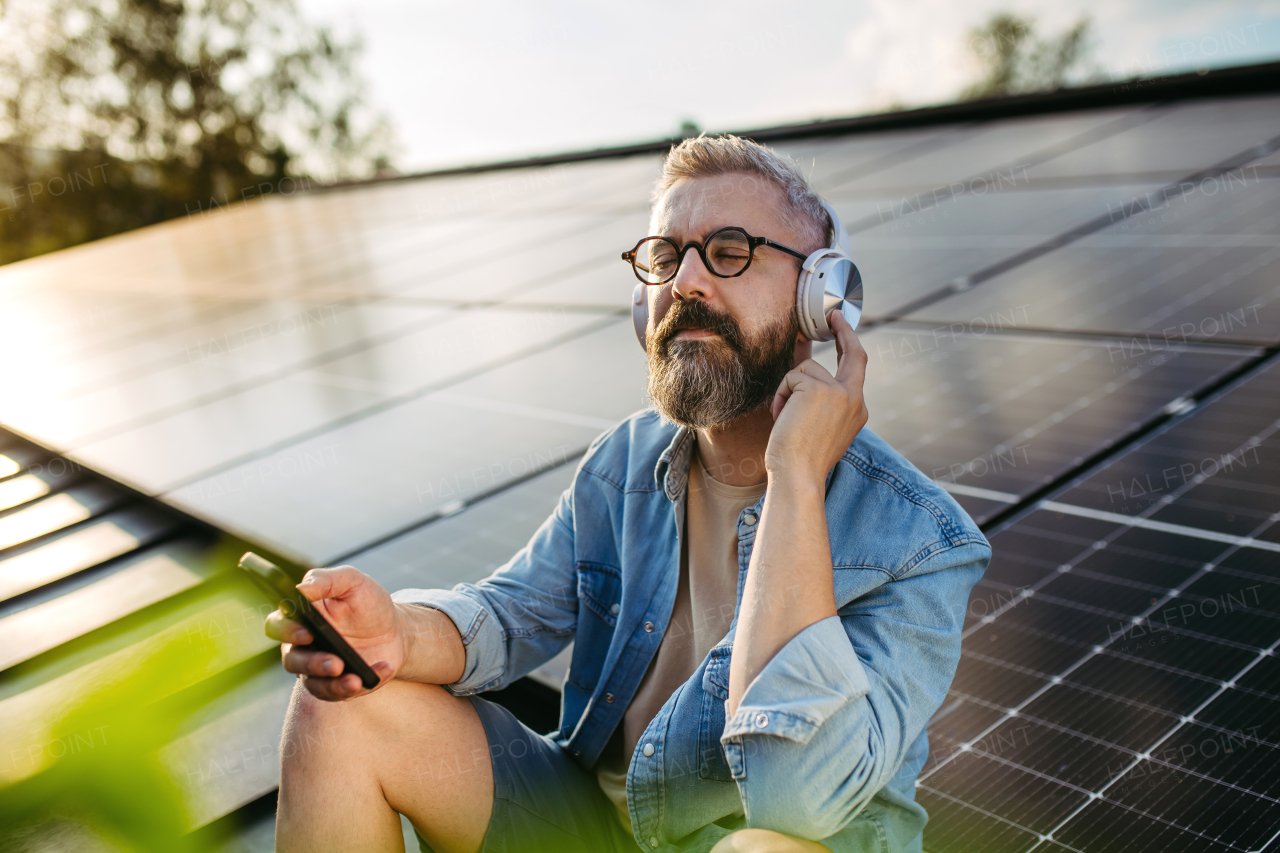 Handsome man sitting on roof full of solar panels, headphones on head, closed eyes.