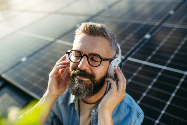 Handsome man sitting on roof full of solar panels, headphones on head, closed eyes.