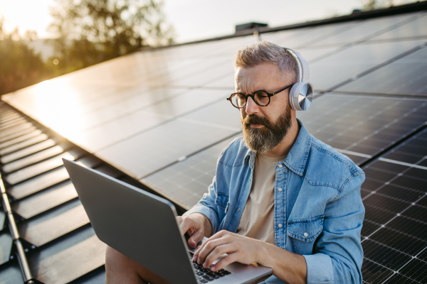 Handsome man sitting on roof full of solar panels, headphones on head, typing on laptop.