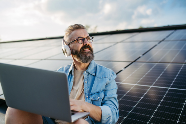 Handsome man sitting on roof full of solar panels, headphones on head, typing on laptop.