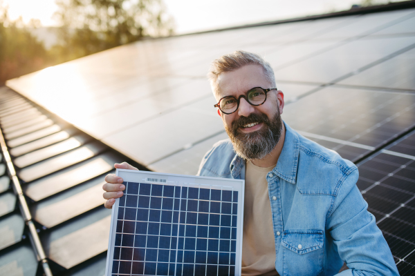 Man on roof with solar panels, looking at camera, holding model of solar panel in arms. Sustainable lifestyle, green household.