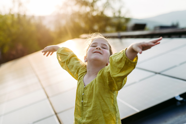 Cute girl on roof with solar panels, standing with open arms, hands up in the air. Rooftop solar or photovoltaic system. Sustainable future for next generation concept.