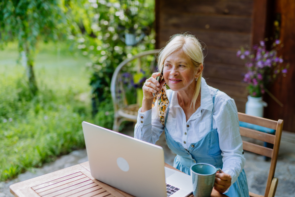 A senior woman with laptop and coffee sitting on terrace in summer, resting.