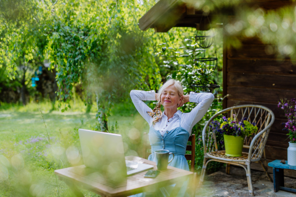 A senior woman with laptop and coffee sitting on terrace in summer, resting.