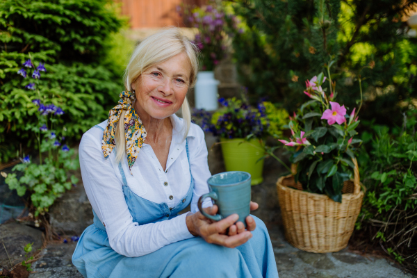 A portrait of senior woman with coffee sitting on terrace in summer, resting and looking at camera.