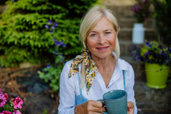 A portrait of senior woman with coffee sitting on terrace in summer, resting and looking at camera.