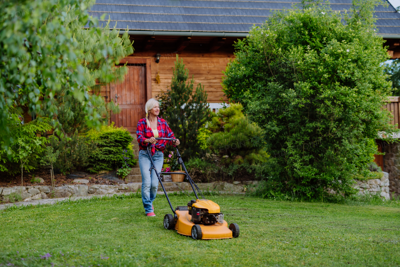 An ederly woman mowing grass with lawn mower in the garden, garden work concept.