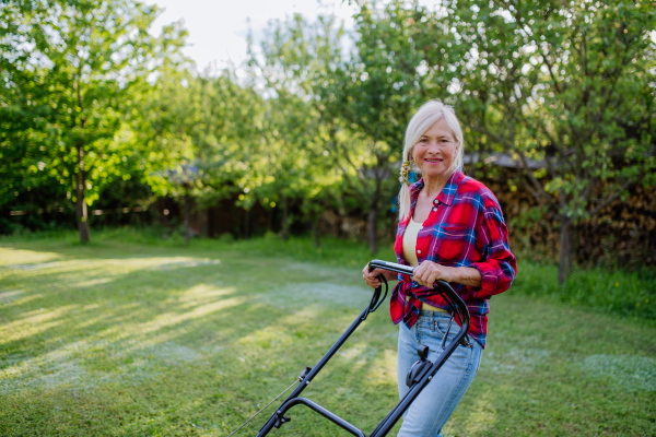 An ederly woman mowing grass with lawn mower in the garden, garden work concept.