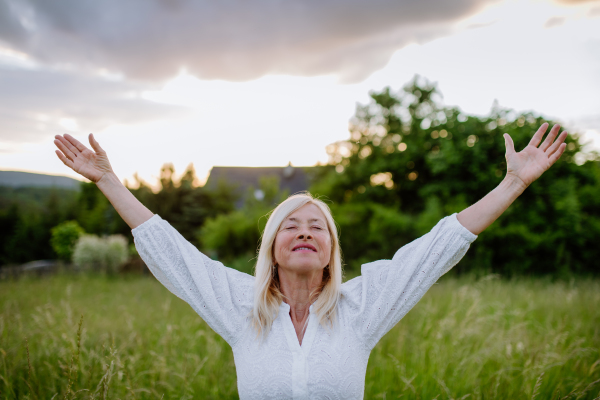 A senior woman with arms outstretched and face up at park on summer day, mental health concept.