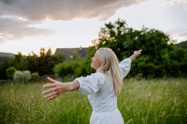 A senior woman with arms outstretched and face up at park on summer day, mental health concept.