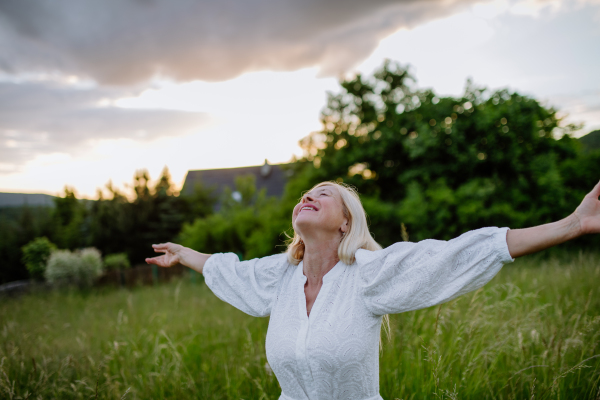 A senior woman with arms outstretched and face up at park on summer day, mental health concept.