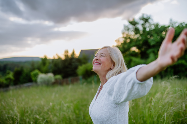 A senior woman with arms outstretched and face up at park on summer day, mental health concept.