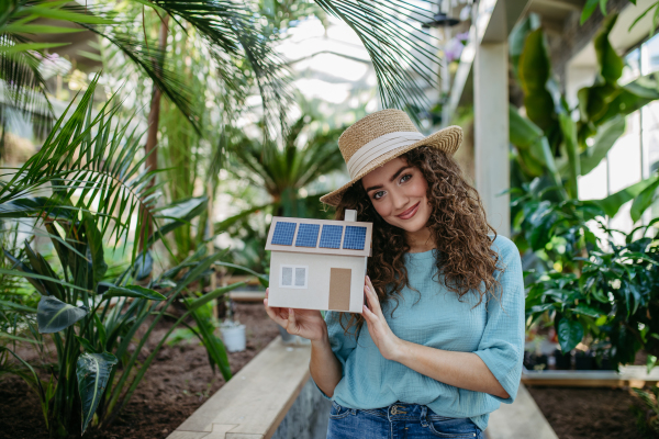 Young woman in a botanical garden holding model of house with solar panels, concept of green energy.