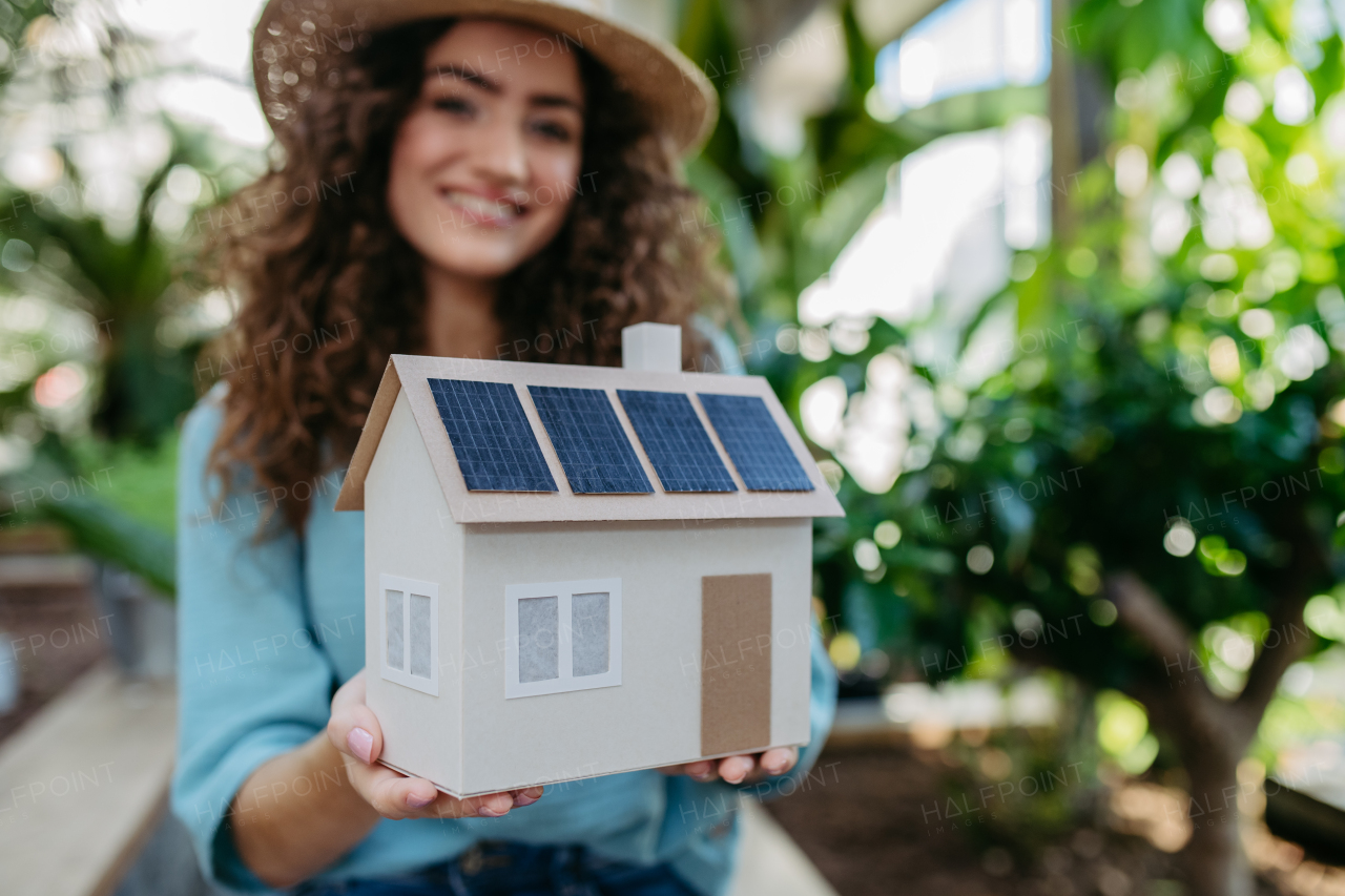 Young woman in a botanical garden holding model of house with solar panels, concept of green energy.