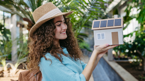 Young woman in a botanical garden holding model of house with solar panels, concept of green energy.