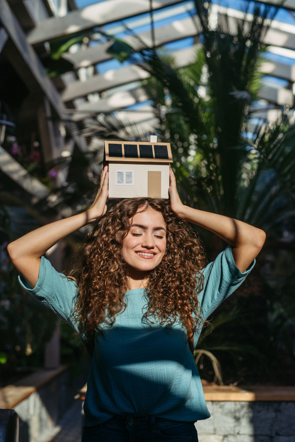 Young woman in a botanical garden holding model of house with solar panels, concept of green energy.