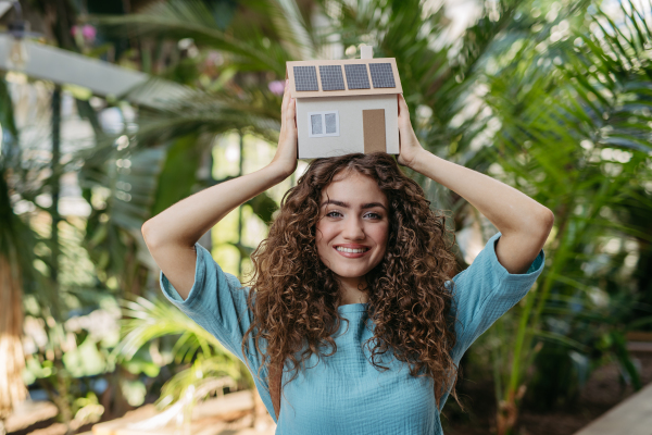 Young woman in a botanical garden holding model of house with solar panels, concept of green energy.