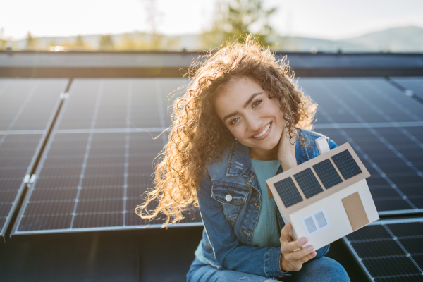 Portrait of young woman, owner on roof with solar panels,holding model of house with photovoltaics.