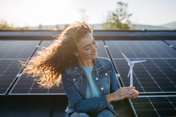 Portrait of young woman, holding model of a wind turbine on roof with solar panels.