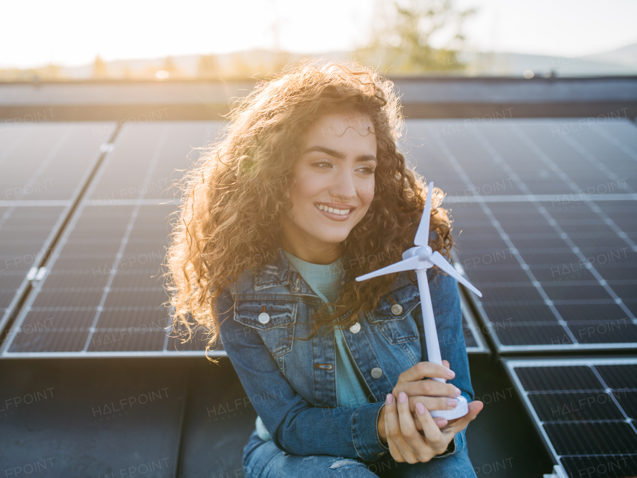 Portrait of young woman, holding model of a wind turbine on roof with solar panels.