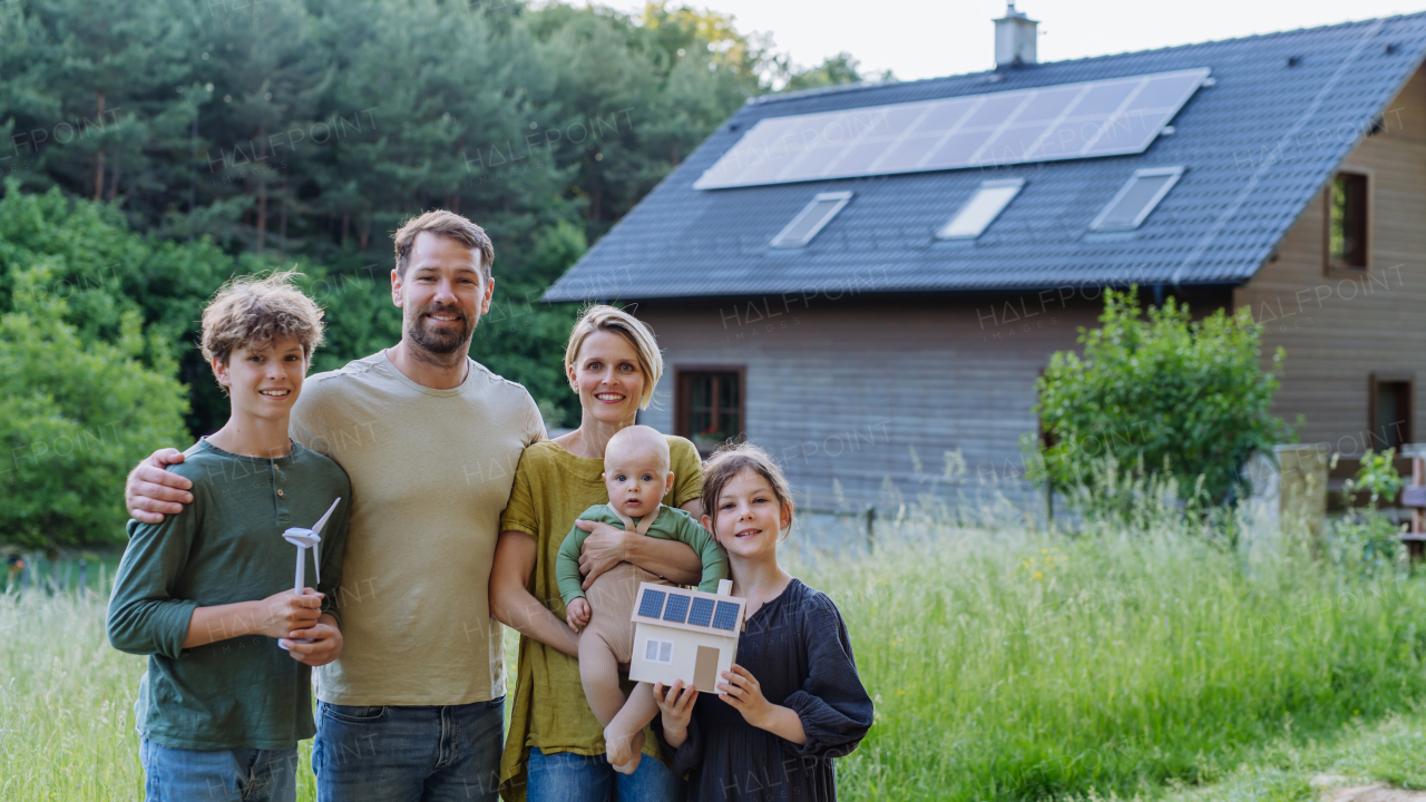 Happy family holding a model of wind turbine and paper house with photovoltaics, standing near their house with solar panels. Alternative energy, saving resources and sustainable lifestyle concept.