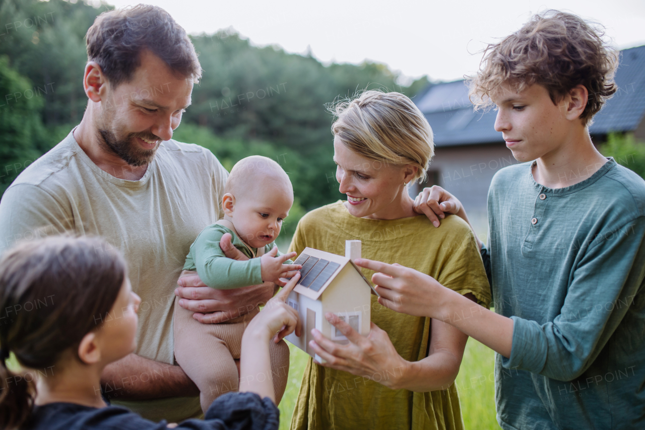 Happy family with three children holding a model of house ith solar photovoltaics. Alternative energy, saving resources and sustainable lifestyle concept.