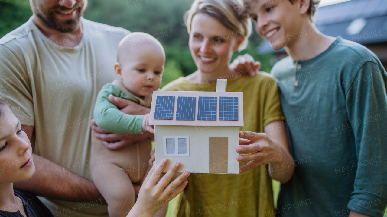 Happy family with three children holding a model of house ith solar photovoltaics. Alternative energy, saving resources and sustainable lifestyle concept.