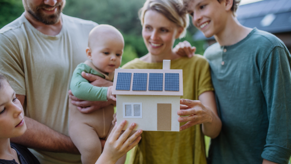 Happy family with three children holding a model of house ith solar photovoltaics. Alternative energy, saving resources and sustainable lifestyle concept.