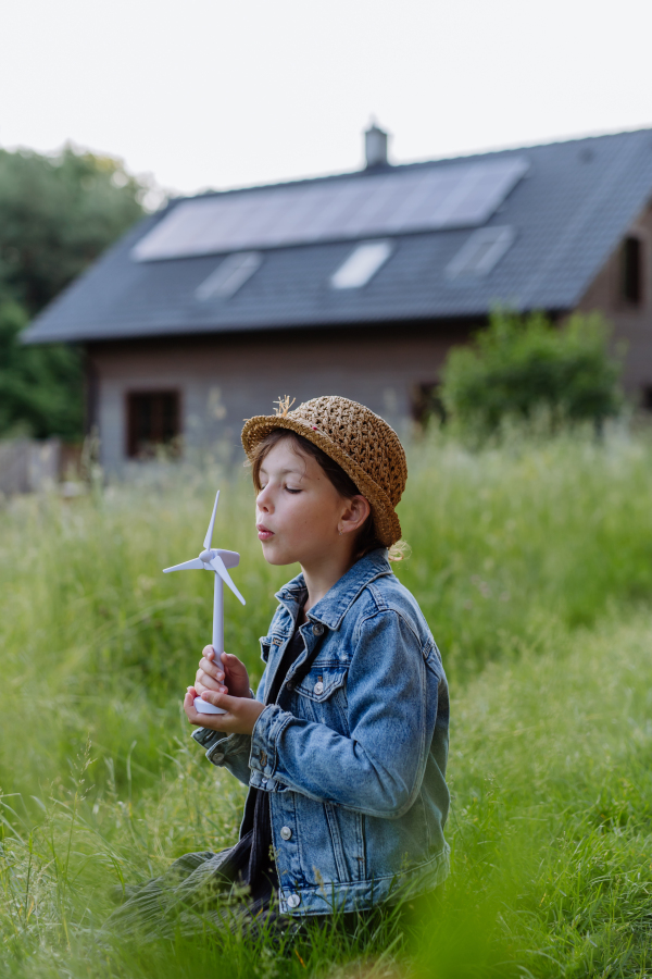 Little girl holding model of wind turbine, standing in front of their house with photovoltaic panels, concept of renewable resources.