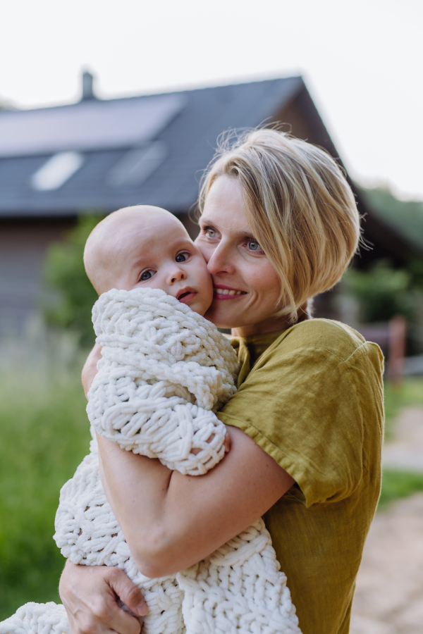 Portrait of a happy mother with her small baby, standing in front of their house.