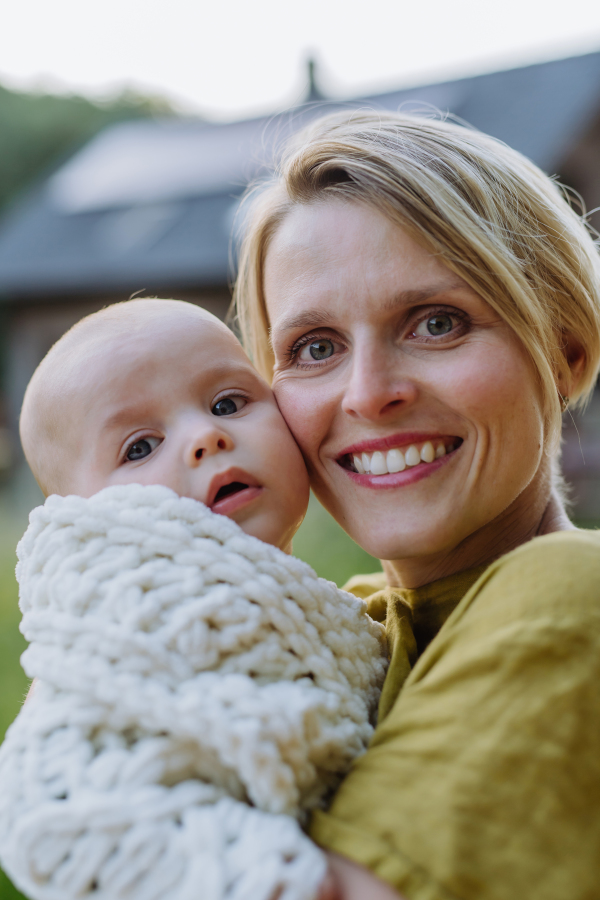 Portrait of a happy mother with her small baby, standing in front of their house.