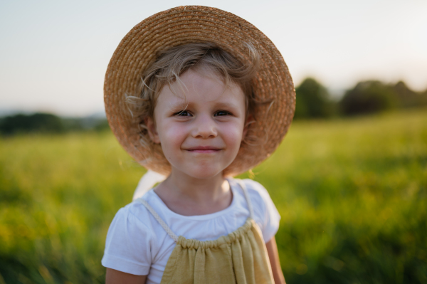 Portrait of adorable little girl in a straw hat standing in the middle of summer meadow. Child with curly blonde hair during sunset. Kids spending summer with grandparents in the countryside.