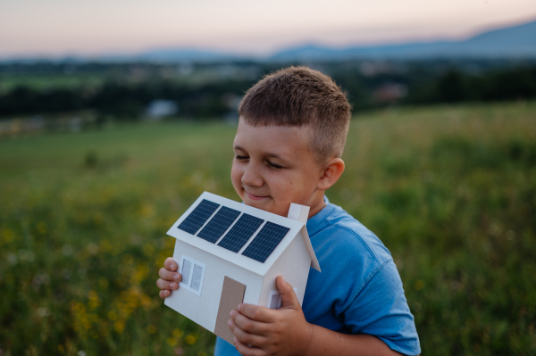 Boy with model house with installed of solar panels, standing in the middle of meadow. Concept of alternative energy, saving resources and sustainable lifestyle concept. . Importance of alternative energy sources and long-term sustainability for future generations