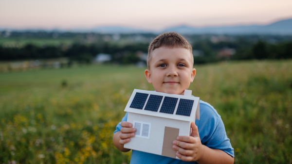 Boy with model house with installed of solar panels, standing in the middle of meadow. Concept of alternative energy, saving resources and sustainable lifestyle concept. . Importance of alternative energy sources and long-term sustainability for future generations