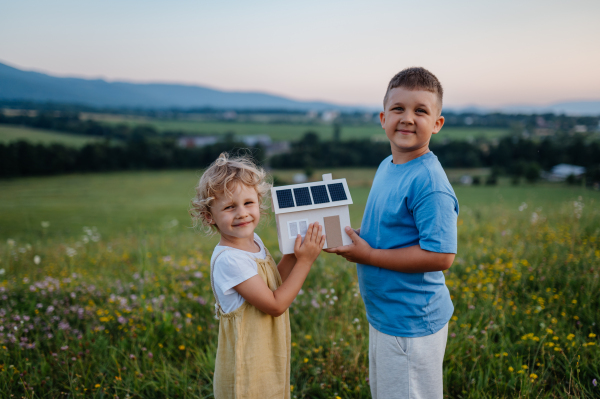 Young boy his sister holding model of house with installed solar panels. Alternative energy, saving resources and sustainable lifestyle concept. Siblings standing in the middle of meadow, representing the importance of green energy for the future generations.