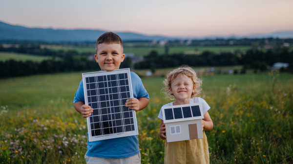 Young boy holding solar panel and his sister holding model of house with installed solar panels. Alternative energy, saving resources and sustainable lifestyle concept. Siblings standing in the middle of meadow, representing the importance of green energy for the future generations.