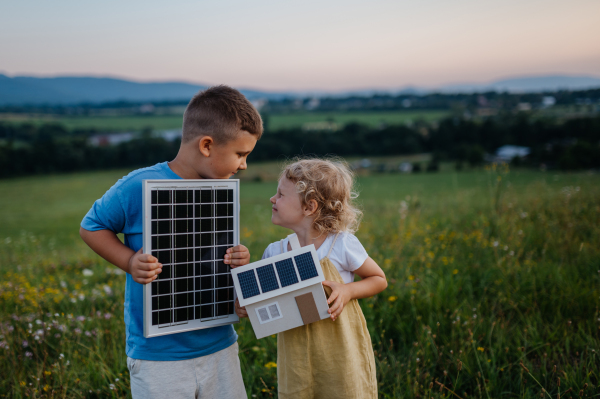 Young boy holding solar panel and his sister holding model of house with installed solar panels. Alternative energy, saving resources and sustainable lifestyle concept. Siblings standing in the middle of meadow, representing the importance of green energy for the future generations.