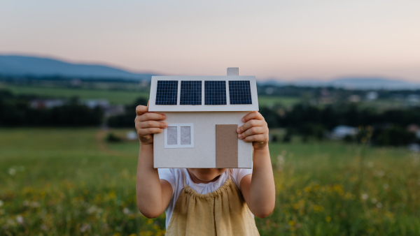 Little girl with model house with installed of solar panels, standing in the middle of meadow. Concept of alternative energy, saving resources and sustainable lifestyle concept. . Importance of alternative energy sources and long-term sustainability for future generations