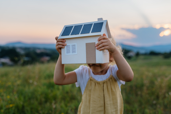 Little girl with model house with installed of solar panels, standing in the middle of meadow. Concept of alternative energy, saving resources and sustainable lifestyle concept. . Importance of alternative energy sources and long-term sustainability for future generations