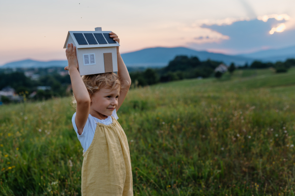 Little girl with model house with installed of solar panels, standing in the middle of meadow. Concept of alternative energy, saving resources and sustainable lifestyle concept. . Importance of alternative energy sources and long-term sustainability for future generations