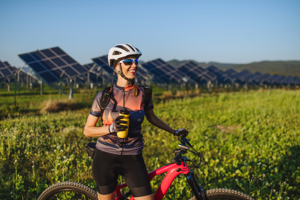 Portrait of a beautiful cyclist standing in front of solar panels at a solar farm, drinking water. A solar farm as solution for more sustainable energy future.